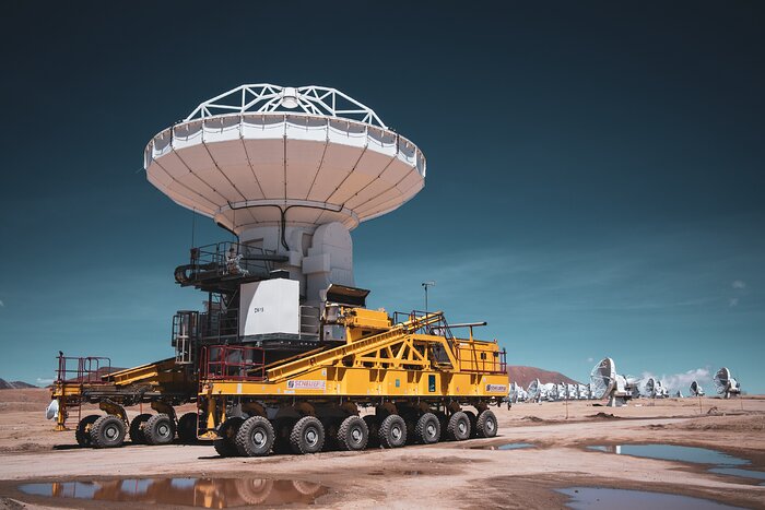 A large white antenna, like a wide cone pointing up, rests on a big orange truck with 28 wheels. There are other similar antennas in the background, on a brown desert landscape under a clear blue sky. Some small ponds of water are present in the foreground.