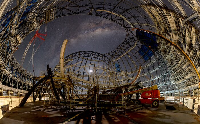 A large metallic structure of intercrossing beams and arches covers most of the image and casts a shadow on the floor in the foreground. On the top left and right corners, sections of the structure are covered by electric-blue panels. The dark-blue night sky is visible through the opening on top of the structure, with the hazy white band of the Milky Way stretching across it.